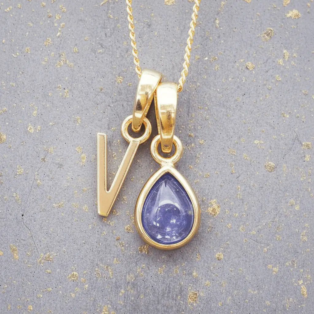 Gold December birthstone necklace with a pear-shaped natural tanzanite pendant and letter 'V' charm with a grey background.