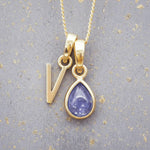 Gold December birthstone necklace with a pear-shaped natural tanzanite pendant and letter 'V' charm with a grey background.