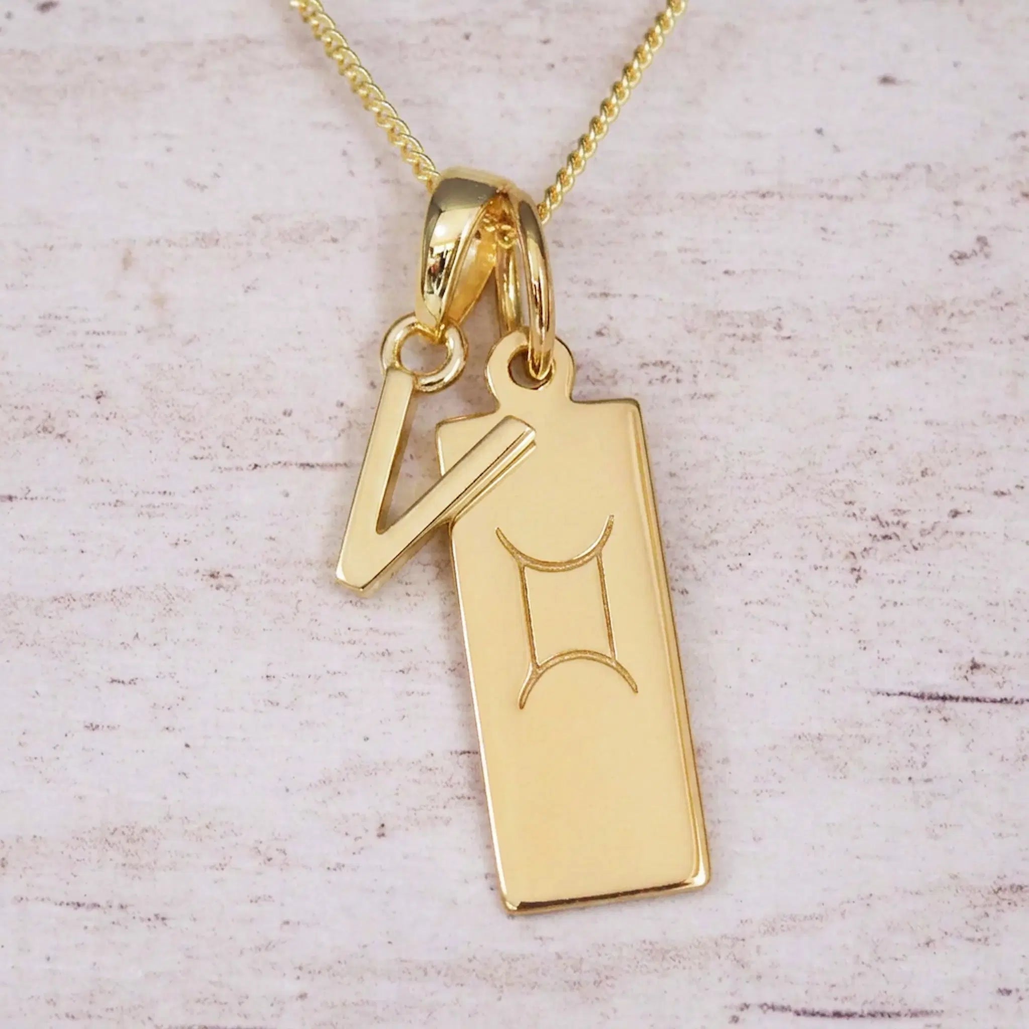 Gold initial V and Gemini zodiac bar necklace, close-up of the letter charm and engraved pendant on whitewashed wood