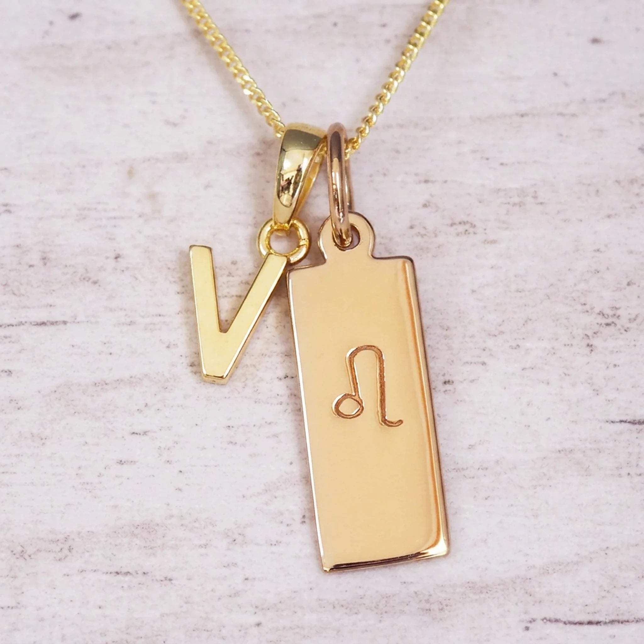 Gold initial and Leo zodiac necklace with a letter V pendant and engraved Leo bar on white wood, close-up