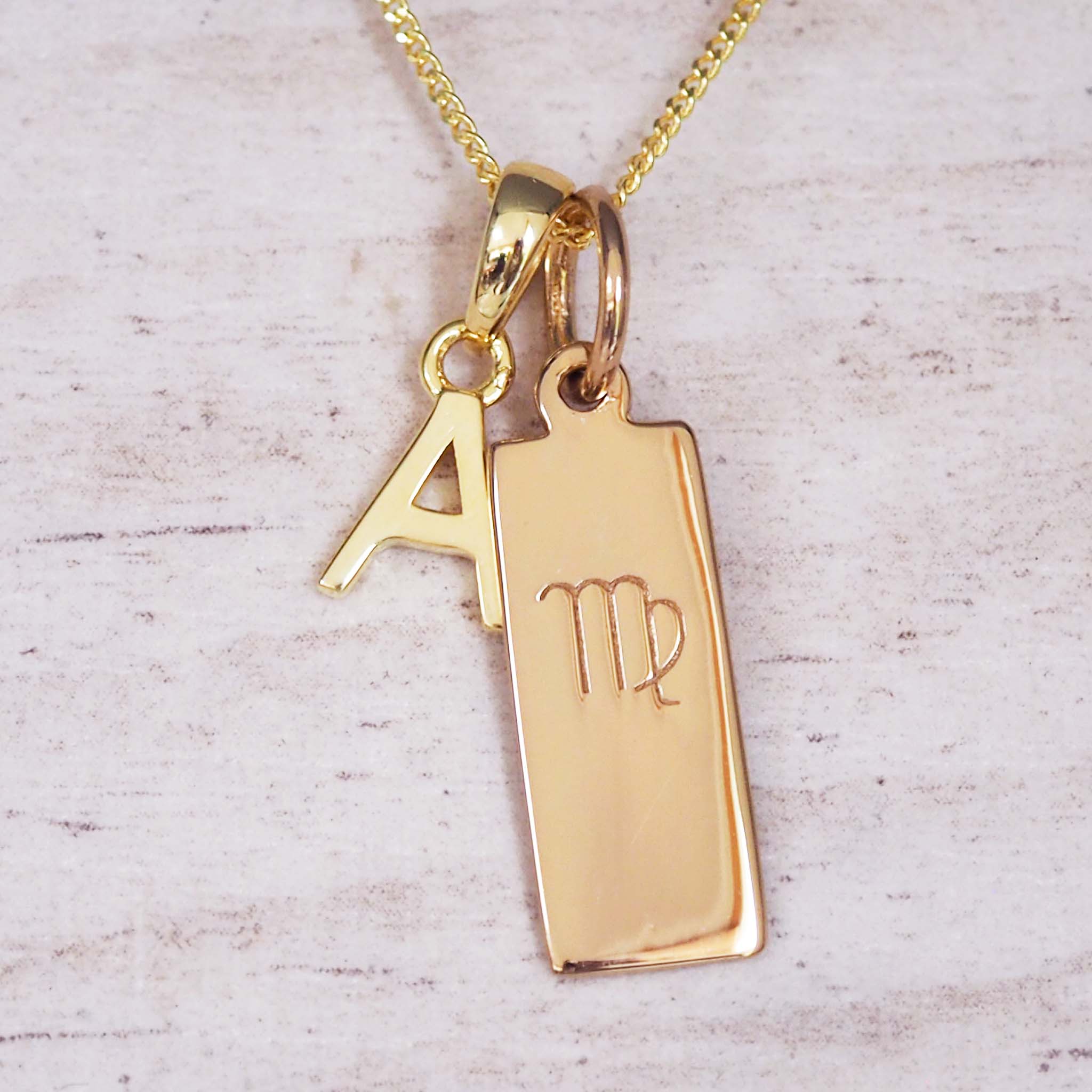 Gold necklace with a pendant featuring the letter 'A' and a virgo zodiac symbol pendant on a light wooden background.
