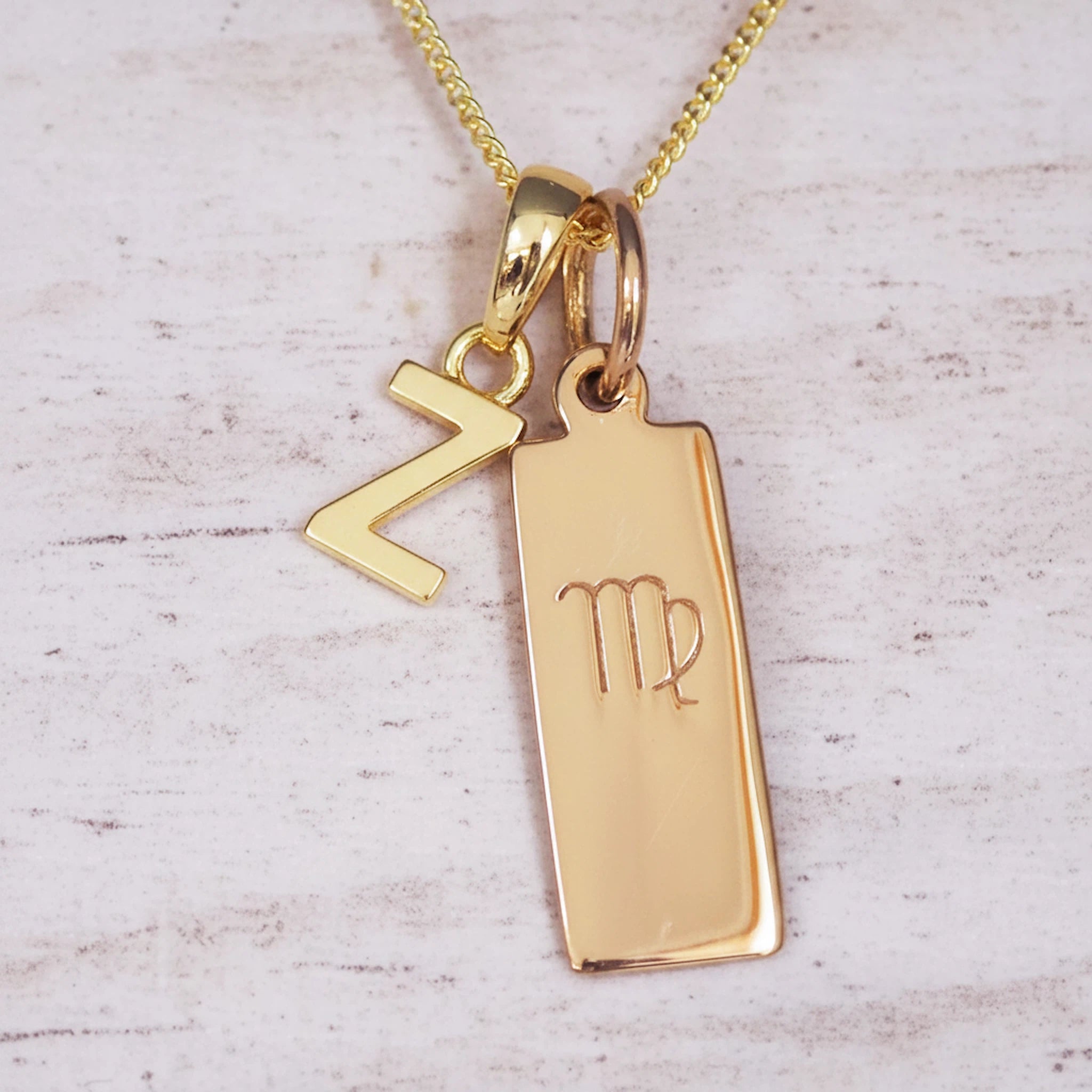 Gold double pendant necklace with a rectangular pendant with virgo zodiac glyph and a letter Z pendant on a light wooden background
