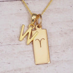 Gold initial W and Aries zodiac bar necklace, close-up of the letter charm and engraved pendant on whitewashed wood