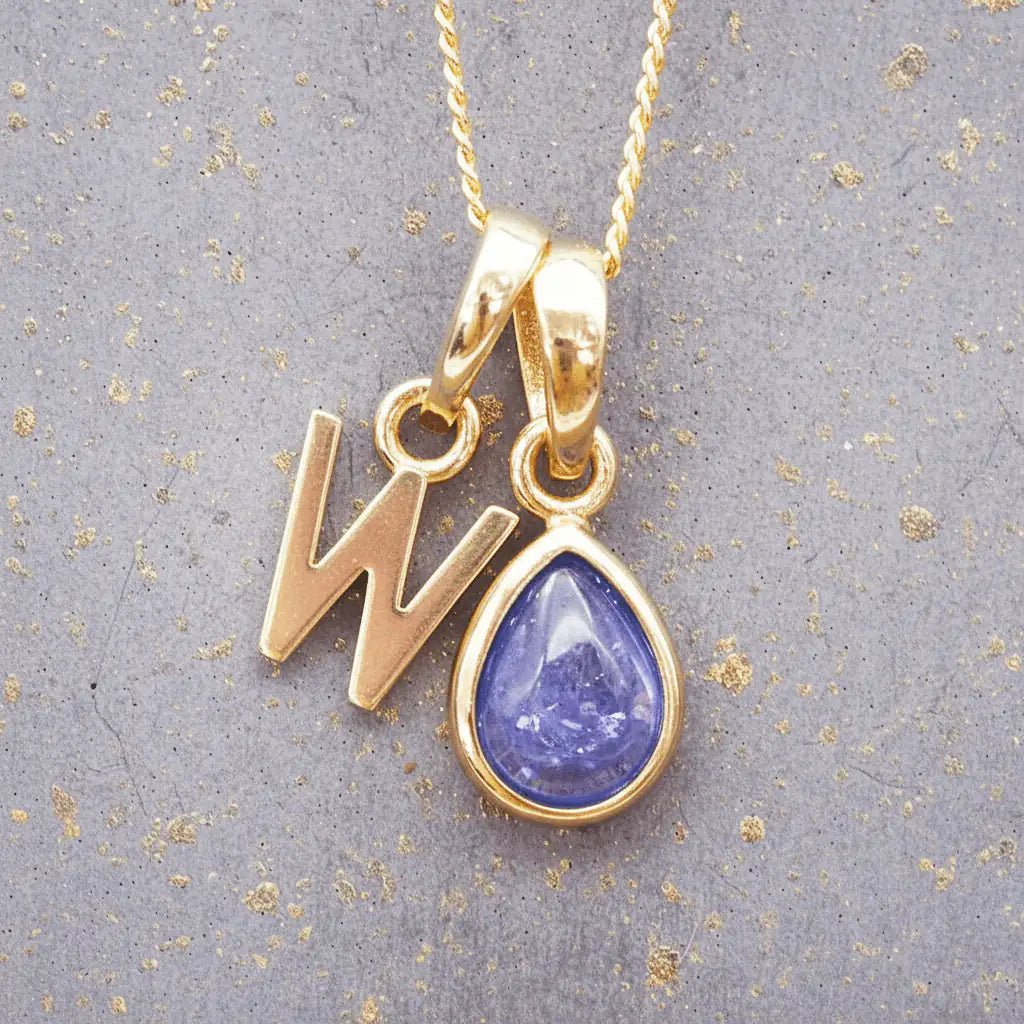 Gold December birthstone necklace with a pear-shaped natural tanzanite pendant and letter 'W' charm with a grey background.
