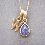 Gold December birthstone necklace with a pear-shaped natural tanzanite pendant and letter 'W' charm with a grey background.