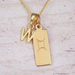 Gold initial W and Gemini zodiac bar necklace, close-up of the letter charm and engraved pendant on whitewashed wood