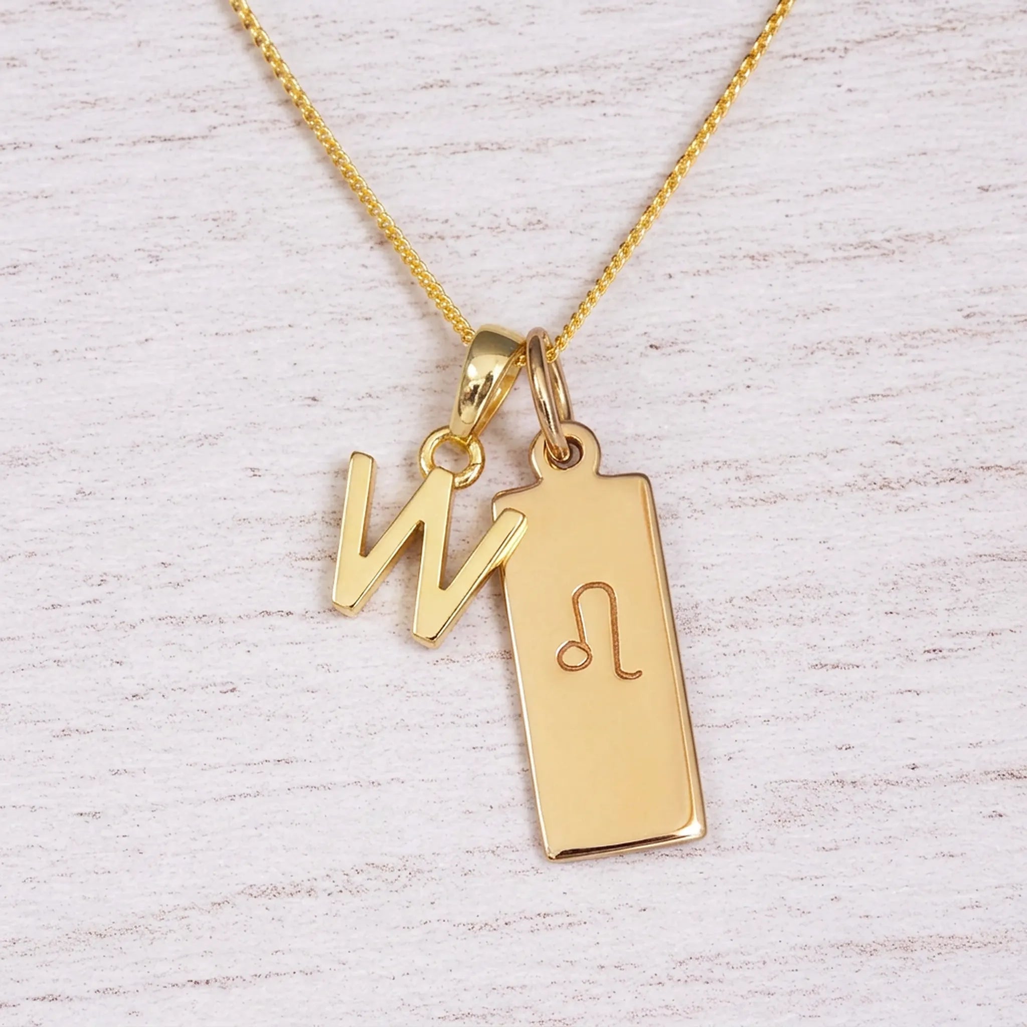 Gold initial and Leo zodiac necklace with a letter W pendant and engraved Leo bar on white wood, close-up