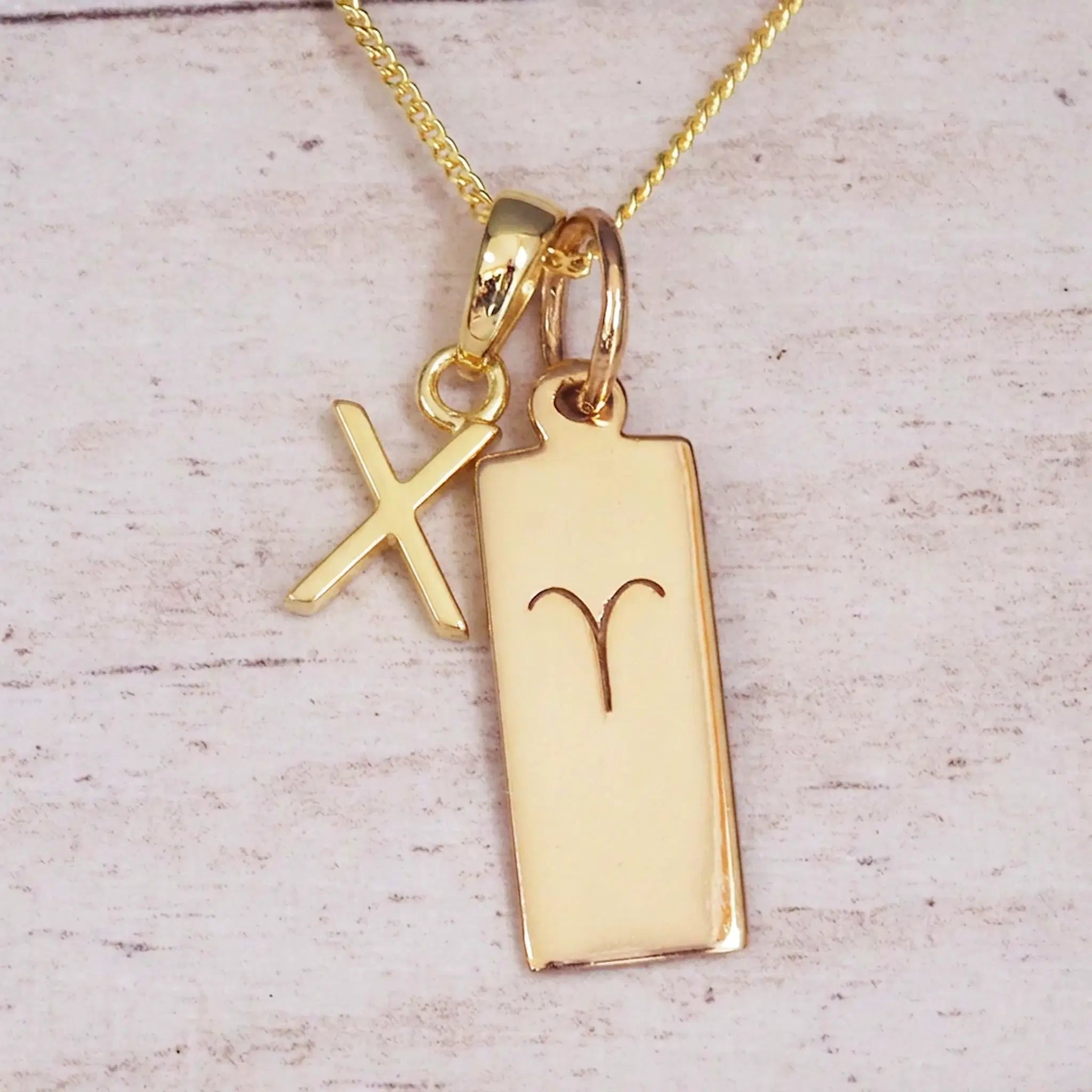 Gold initial X and Aries zodiac bar necklace, close-up of the letter charm and engraved pendant on whitewashed wood