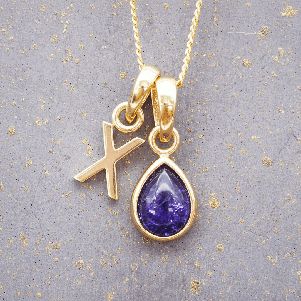 Gold necklace with a purple teardrop december birthstone tanzanite pendant and 'X' initial charm on a grey background