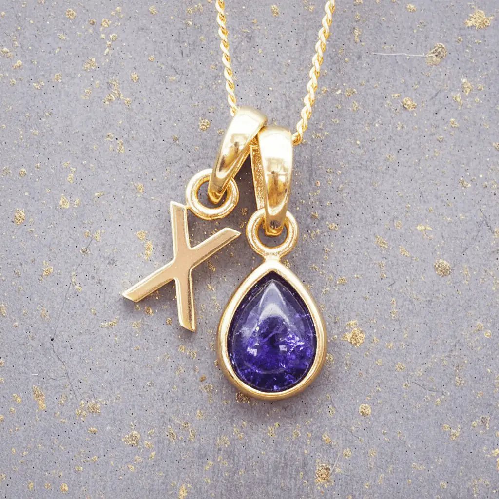 Gold December birthstone necklace with a pear-shaped natural tanzanite pendant and letter 'X' charm with a grey background.