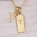 Gold initial Y and Aries zodiac bar necklace, close-up of the letter charm and engraved pendant on whitewashed wood