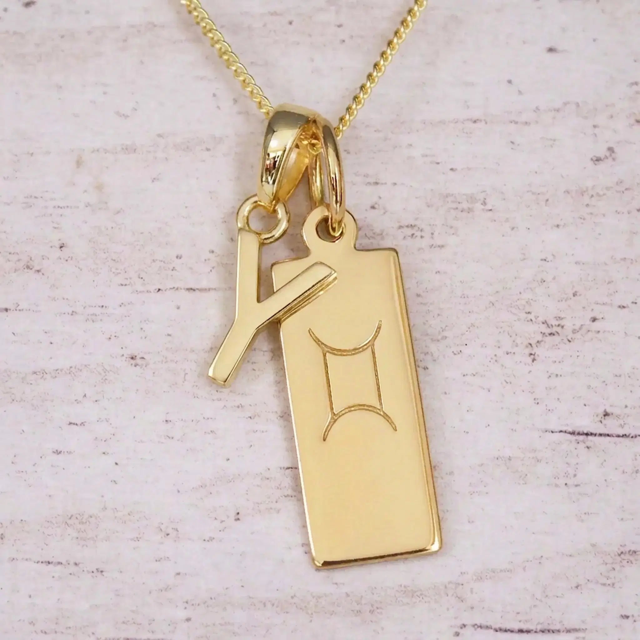 Gold initial Y and Gemini zodiac bar necklace, close-up of the letter charm and engraved pendant on whitewashed wood