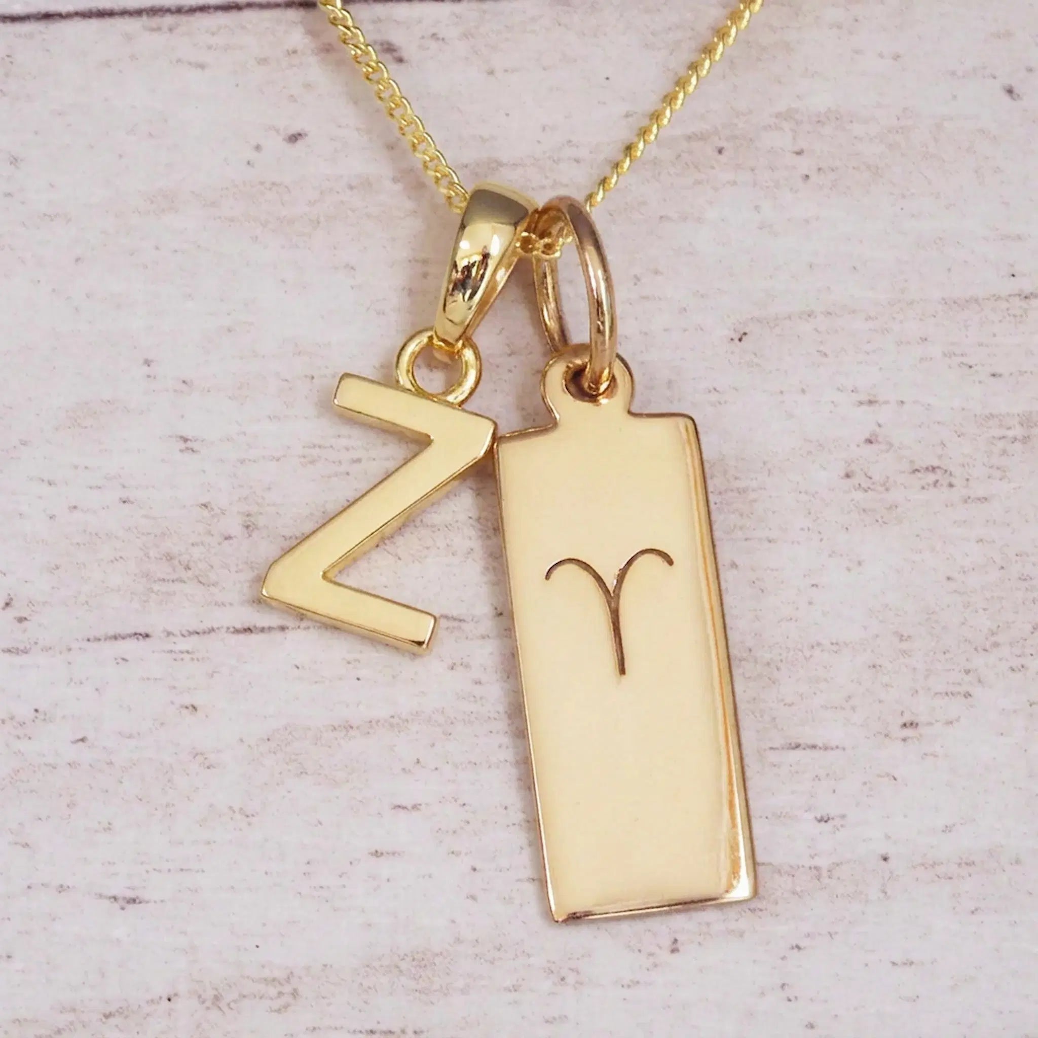 Gold initial Z and Aries zodiac bar necklace, close-up of the letter charm and engraved pendant on whitewashed wood