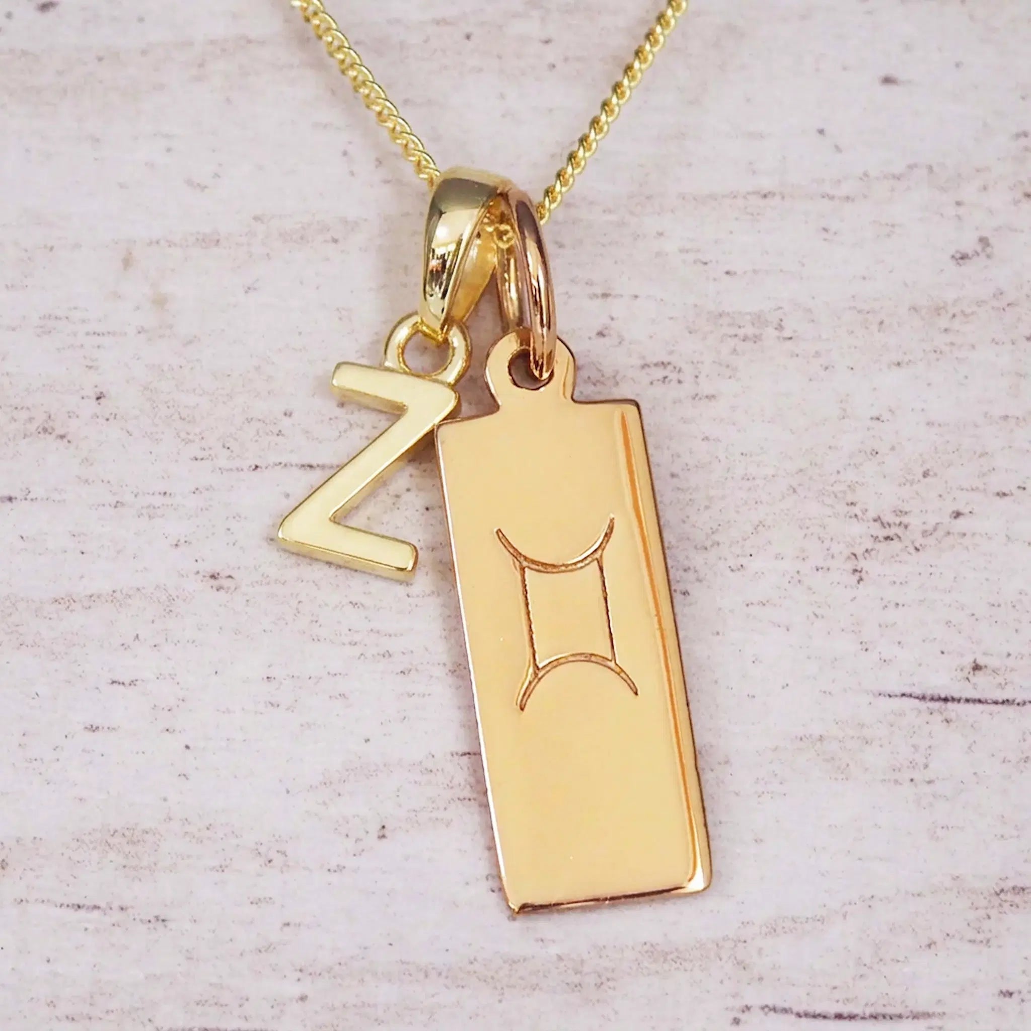 Gold initial Z and Gemini zodiac bar necklace, close-up of the letter charm and engraved pendant on whitewashed wood