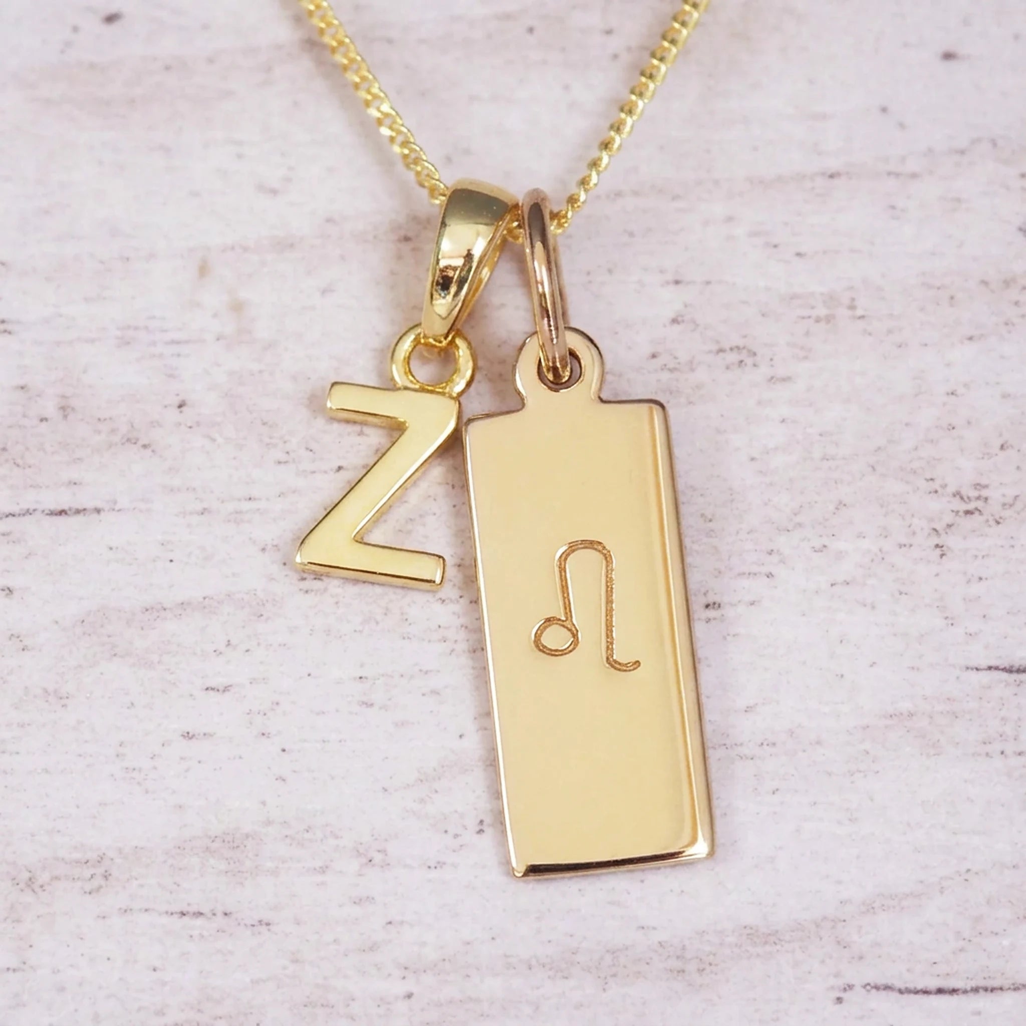 Gold initial and Leo zodiac necklace with a letter Z pendant and engraved Leo bar on white wood, close-up