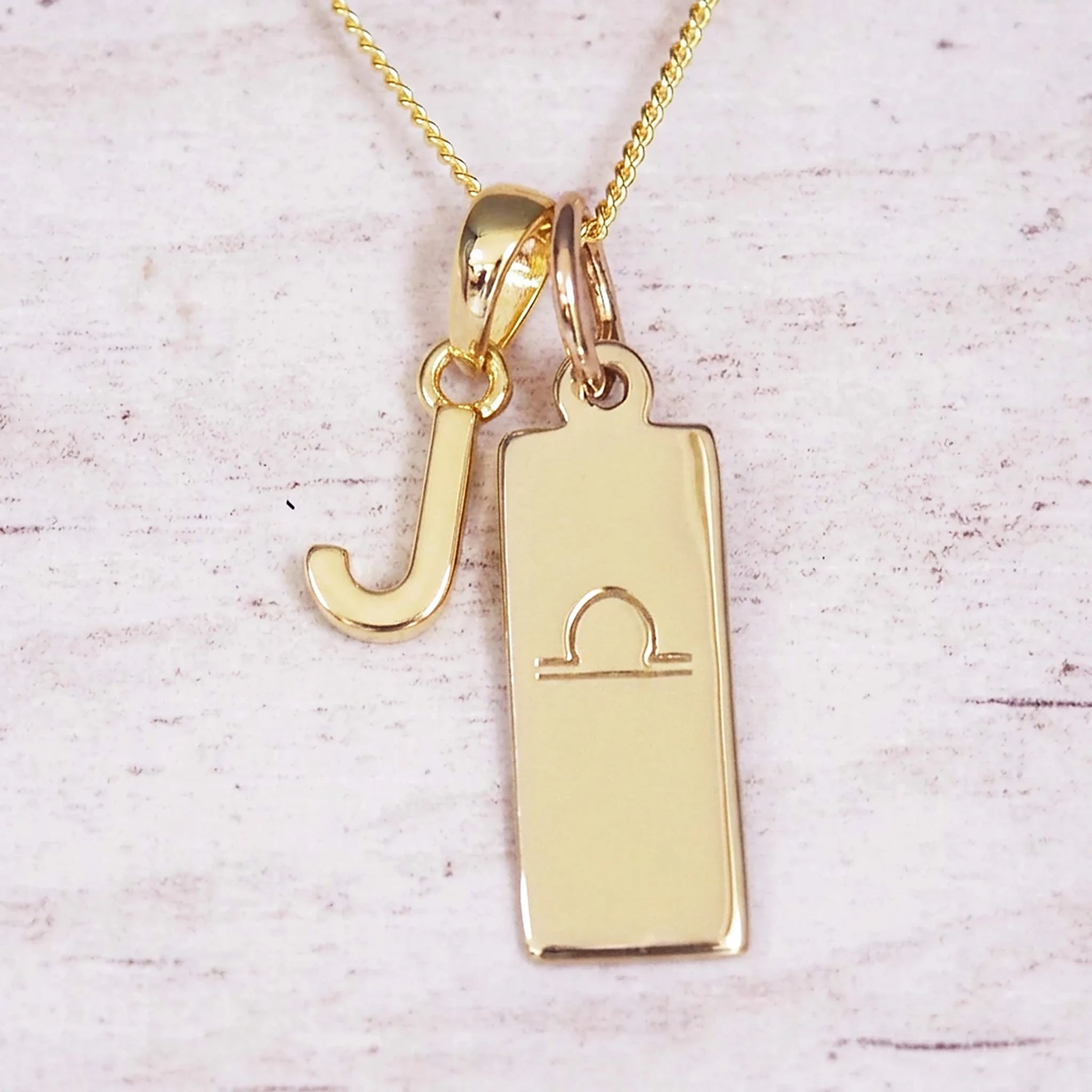 Gold initial and Libra zodiac necklace with a letter J charm and engraved bar pendant on whitewashed wood