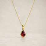 gold chain necklace with a january birthstone teardrop-shaped dark red natural garnet gemstone pendant, displayed against a neutral textured beige background.