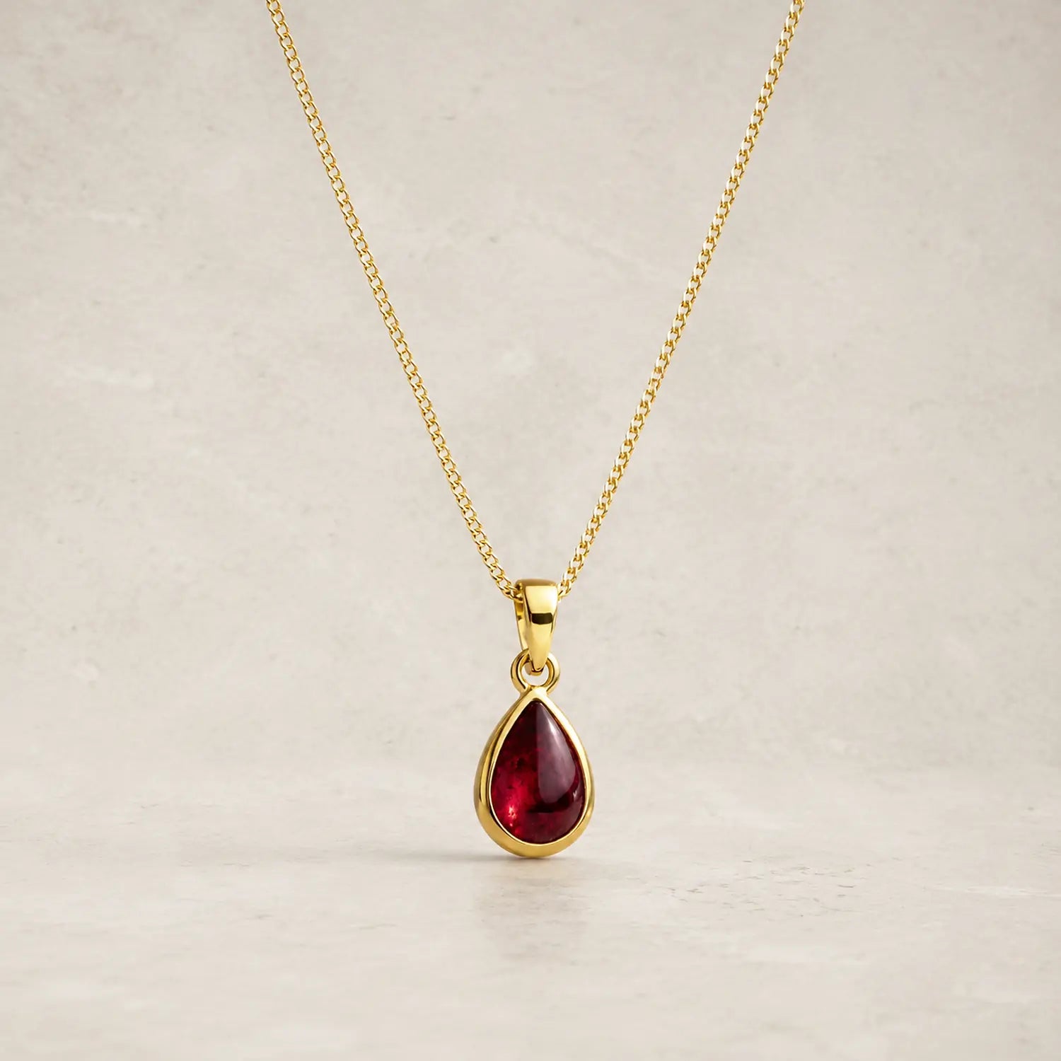 gold chain necklace with a january birthstone teardrop-shaped dark red natural garnet gemstone pendant, displayed against a neutral textured beige background.