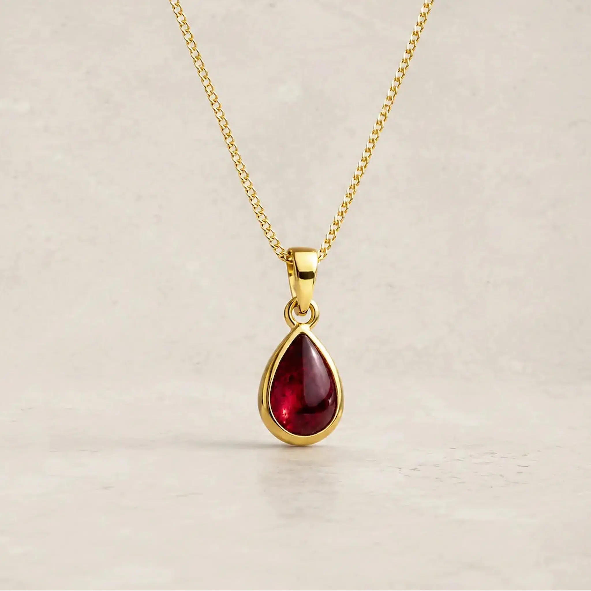 gold chain necklace with a january birthstone teardrop-shaped dark red natural garnet gemstone pendant, displayed against a neutral textured beige background.