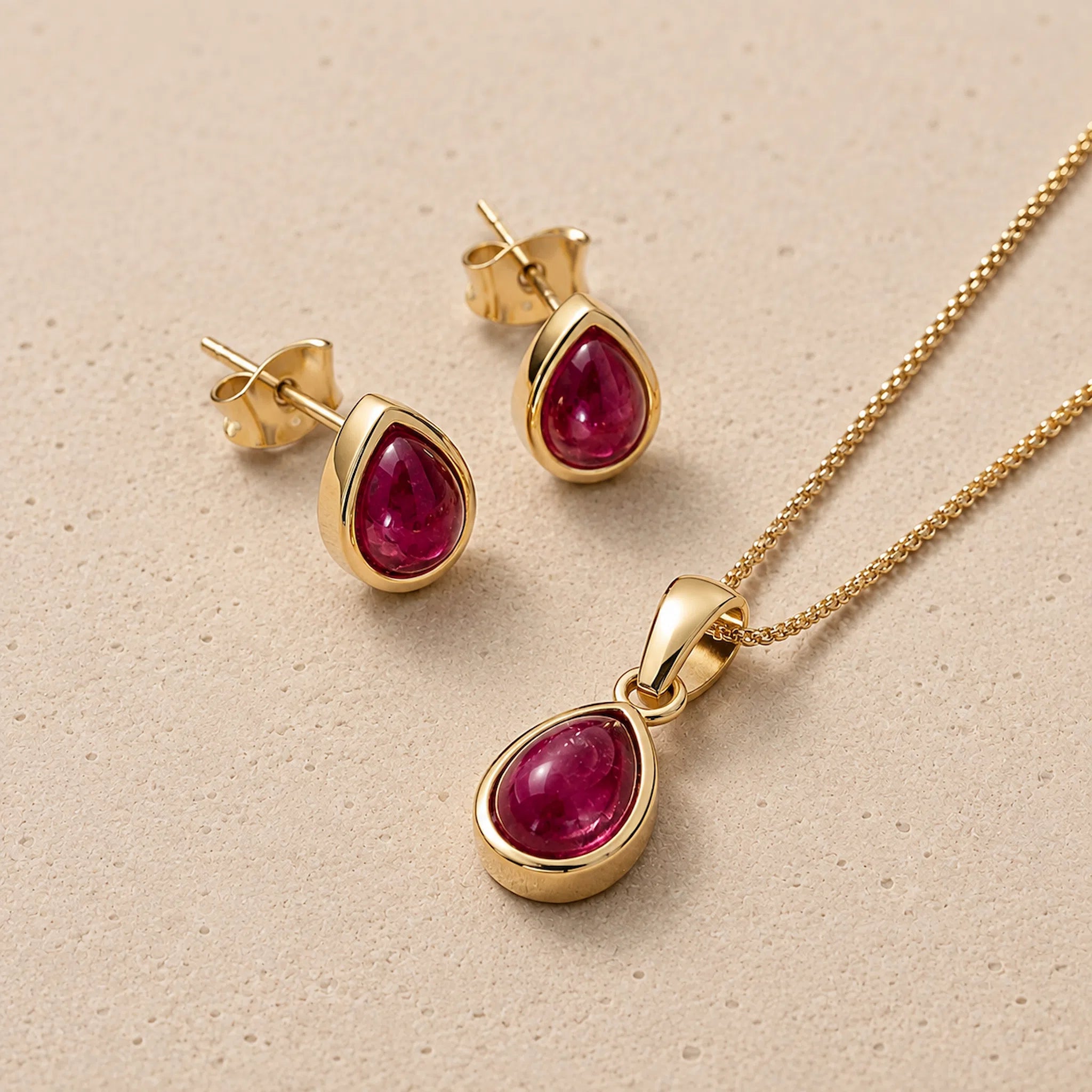 Gold July Birthstone Jewellery set featuring a teardrop-shaped pendant necklace and matching stud earrings with deep red natural ruby gemstones, displayed on a textured beige surface.
