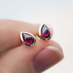A pair of july birthstone gold teardrop stud earrings with deep red natural ruby gemstones, shown worn on a person's finger against a soft white background.