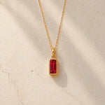 Gold fine chain necklace with a rectangle faceted bezel set july cubic zirconia pendant on a light stone background.