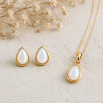 Gold June Birthstone Jewellery set featuring a teardrop pendant pearl necklace and matching stud pearl earrings with natural milky white teardrop pearls, arranged on a textured beige surface with dried flowers.