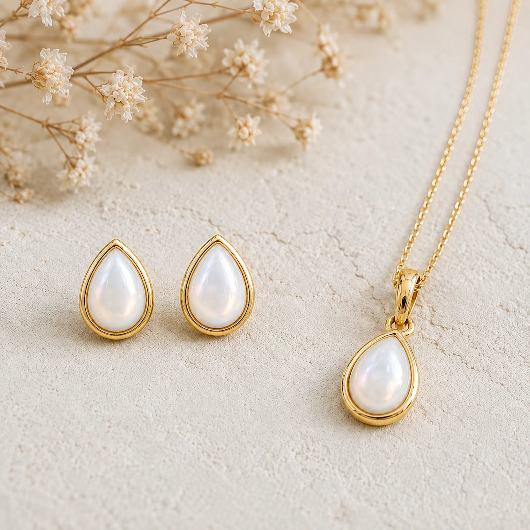 Gold June Birthstone Jewellery set featuring a teardrop pendant pearl necklace and matching stud pearl earrings with natural milky white teardrop pearls, arranged on a textured beige surface with dried flowers.