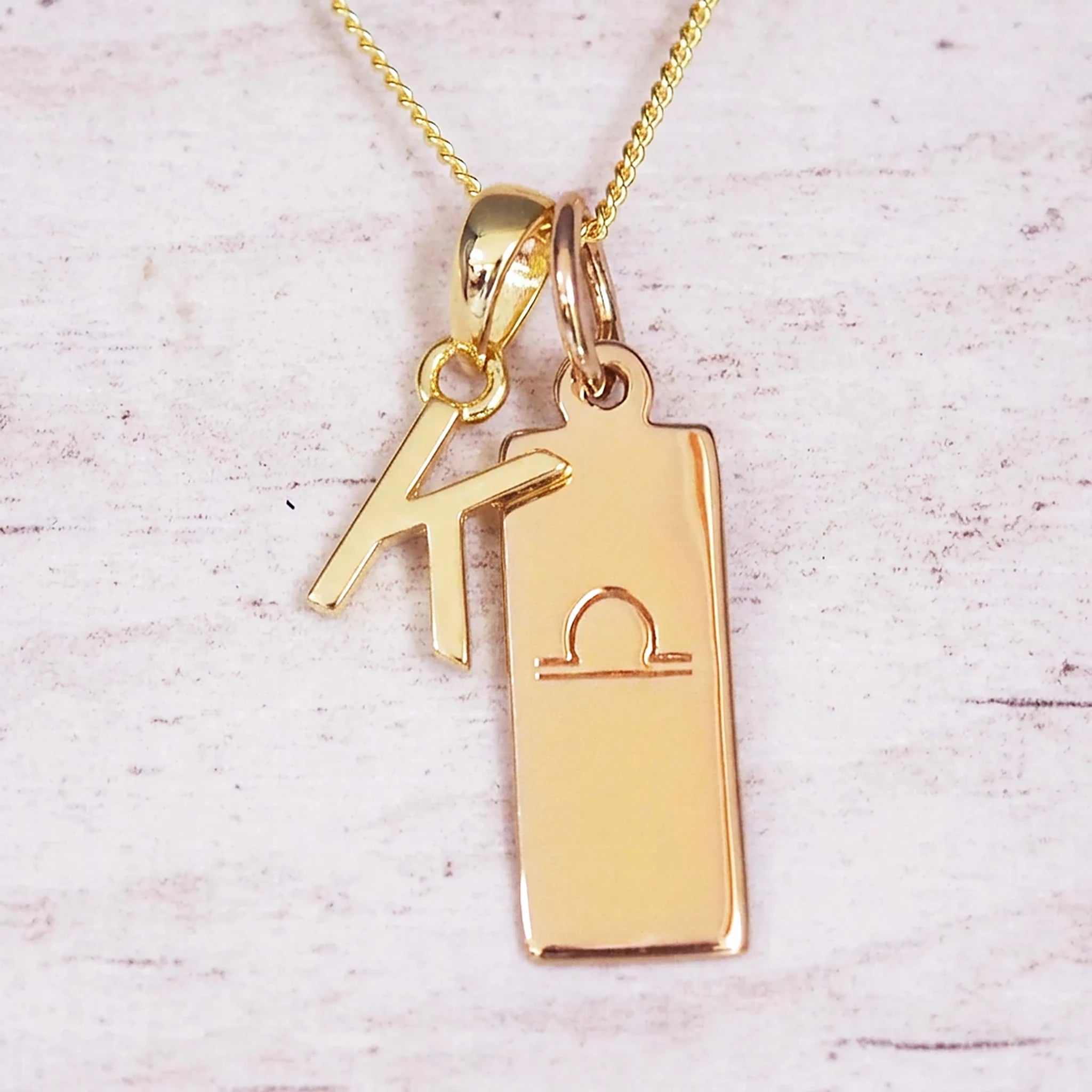 Gold initial and Libra zodiac necklace with a letter K charm and engraved bar pendant on whitewashed wood