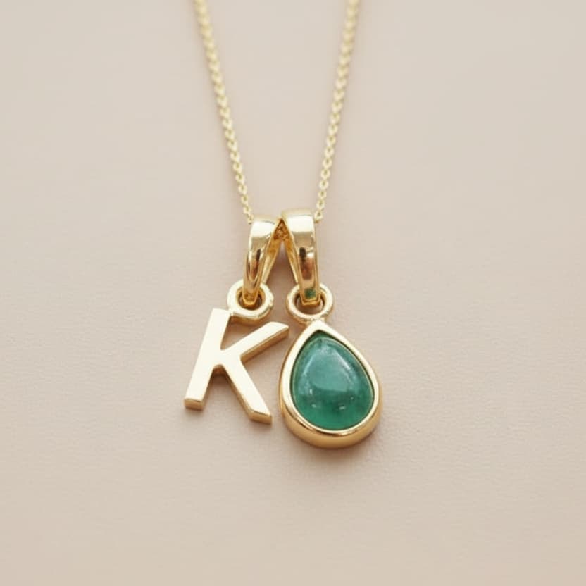 Gold necklace with 'K' pendant and green teardrop charm on a beige background