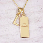 Gold initial and Libra zodiac necklace with a letter L charm and engraved bar pendant on whitewashed wood