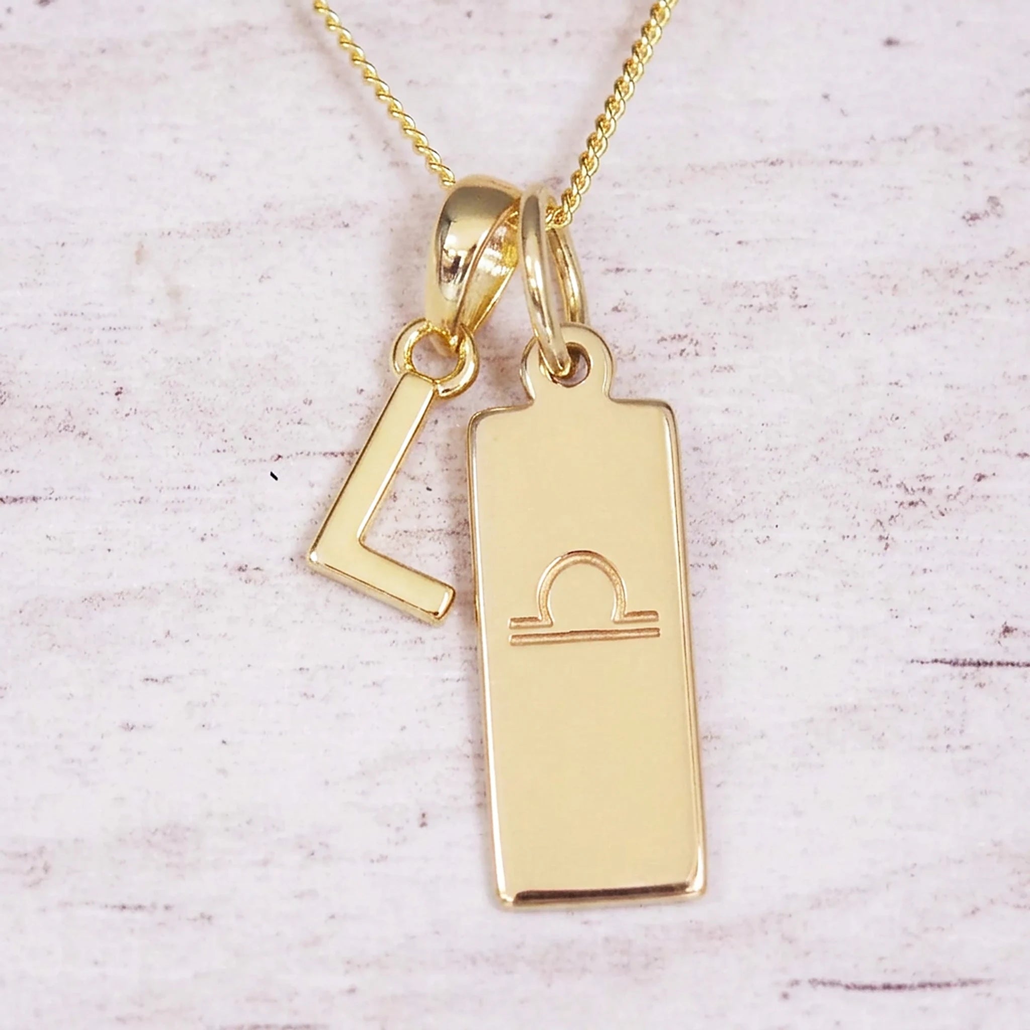 Gold initial and Libra zodiac necklace with a letter L charm and engraved bar pendant on whitewashed wood