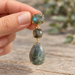 Gold labradorite drop earring held between fingers against an earthy background, showing natural stone details.