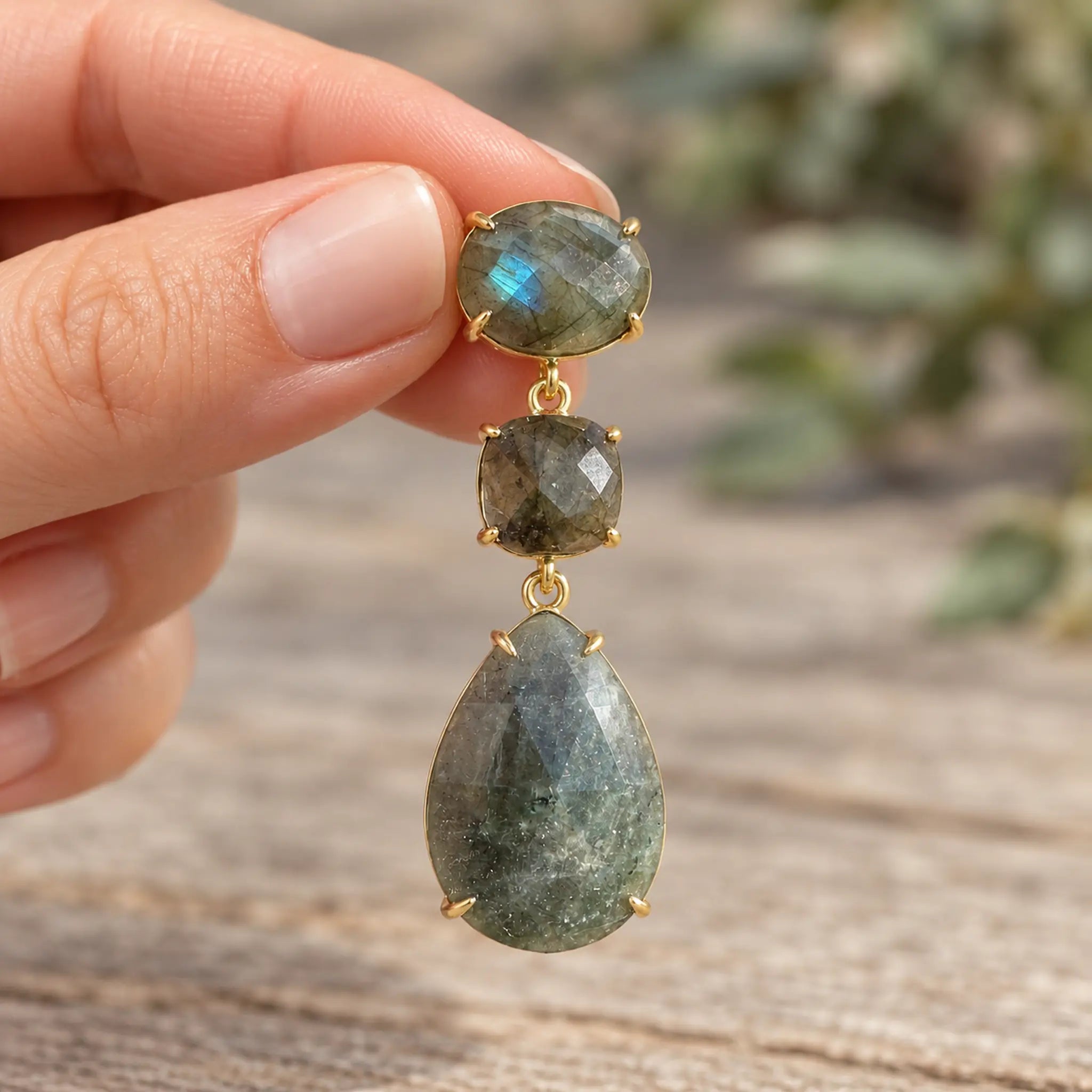 Gold labradorite drop earring held between fingers against an earthy background, showing natural stone details.