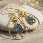 A pair of gold teardrop dangle earrings with blue labradorite stones, displayed on a white stone over a light wood surface.