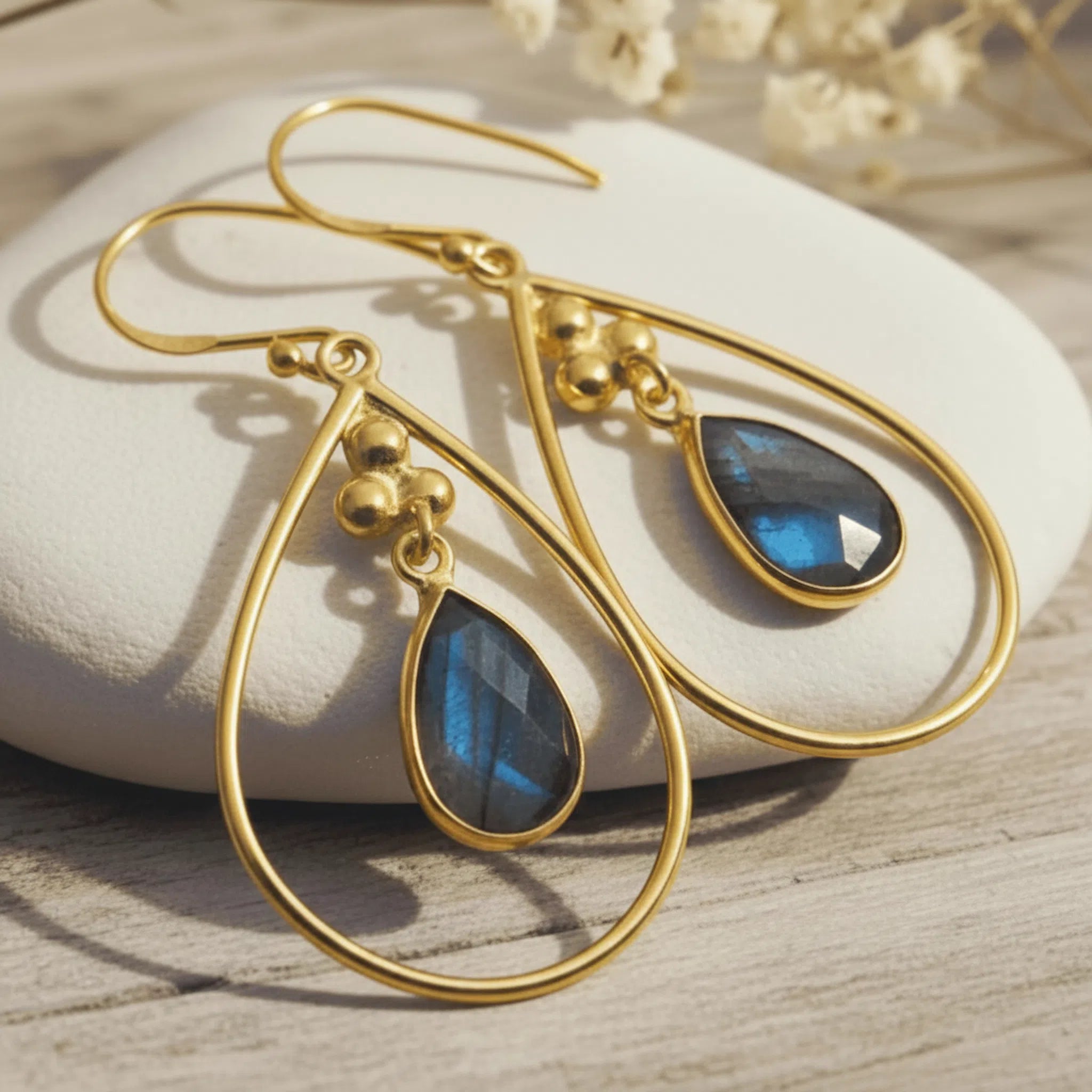 A pair of gold teardrop dangle earrings with blue labradorite stones, displayed on a white stone over a light wood surface.
