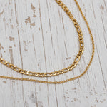 Two gold-tone chain necklaces, one thick curb chain and one delicate cable chain, displayed on a distressed white wooden surface.
