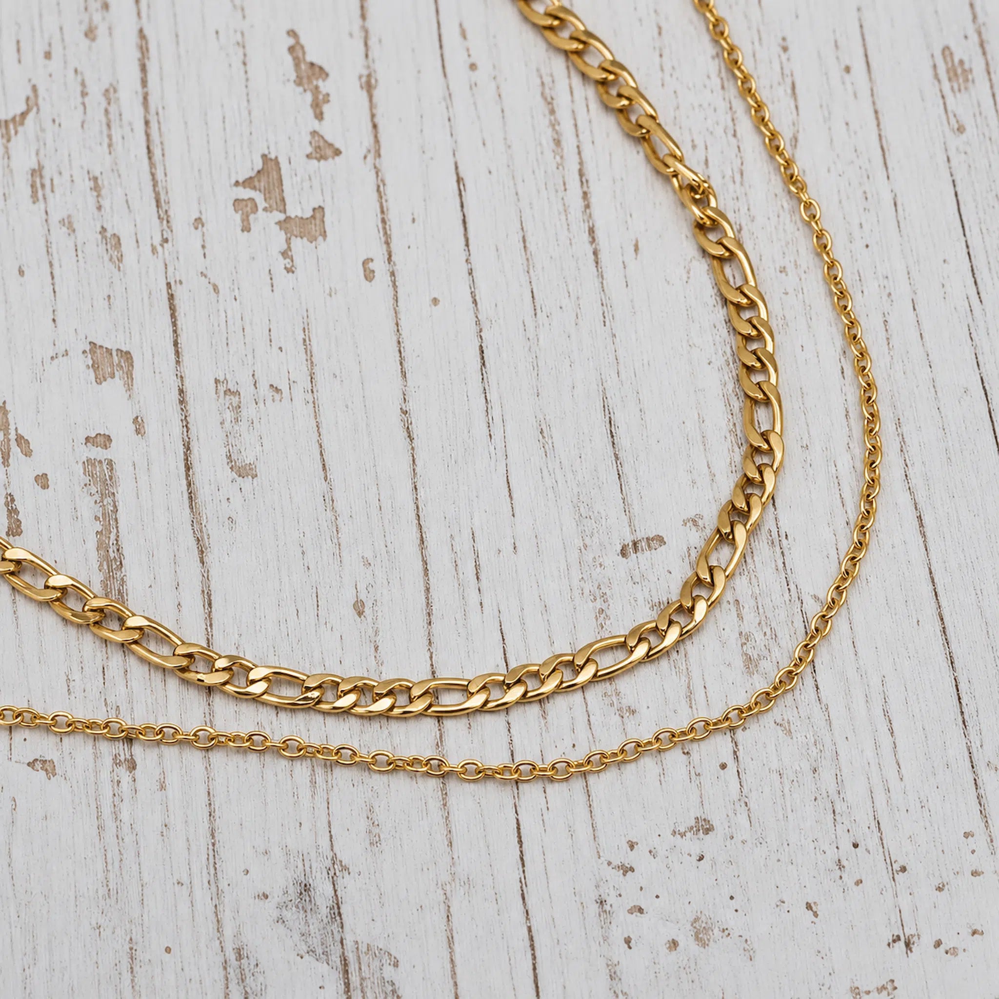 Two gold-tone chain necklaces, one thick curb chain and one delicate cable chain, displayed on a distressed white wooden surface.