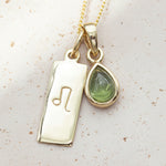 Gold necklace with a green teardrop natural peridot pendant and a rectangular leo zodiac pendant on a textured surface