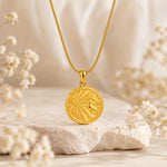 A gold pendant necklace with a circular leo zodiac sign design, resting on a textured stone with soft dried flowers in a warm, neutral setting.