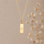 Gold necklace with a rectangular pendant featuring a libra zodiac symbol on a beige background.