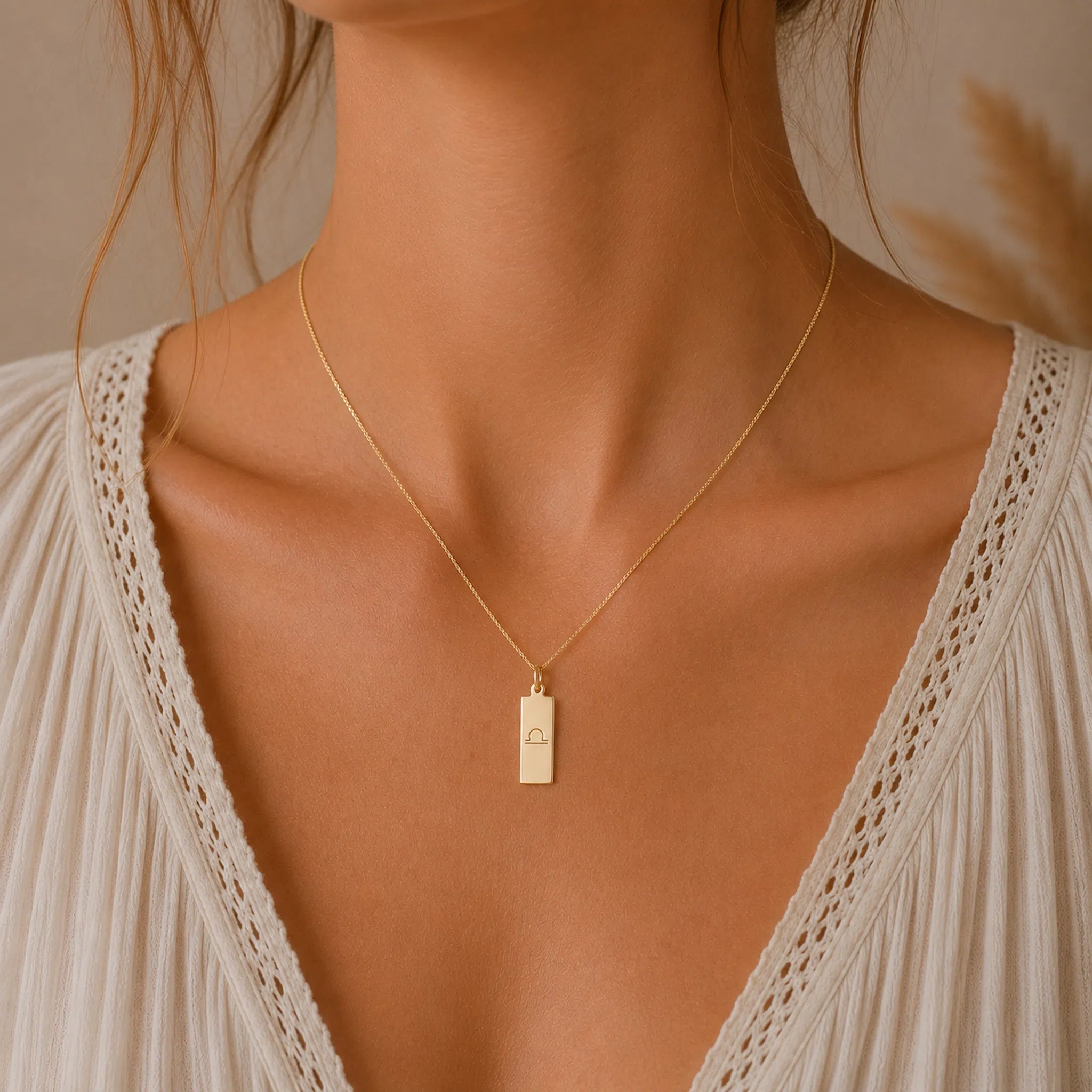 Gold necklace with a libra line glyph pendant on a person wearing a white lace top against a neutral background