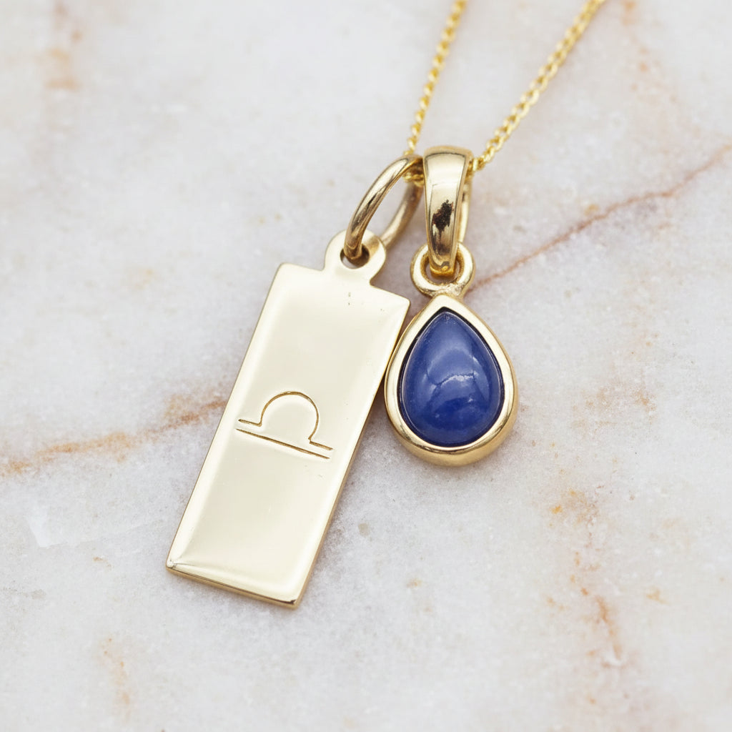 Gold necklace with a blue teardrop natural sapphire gemstone pendant and a gold libra zodiac pendant on a warm stone background