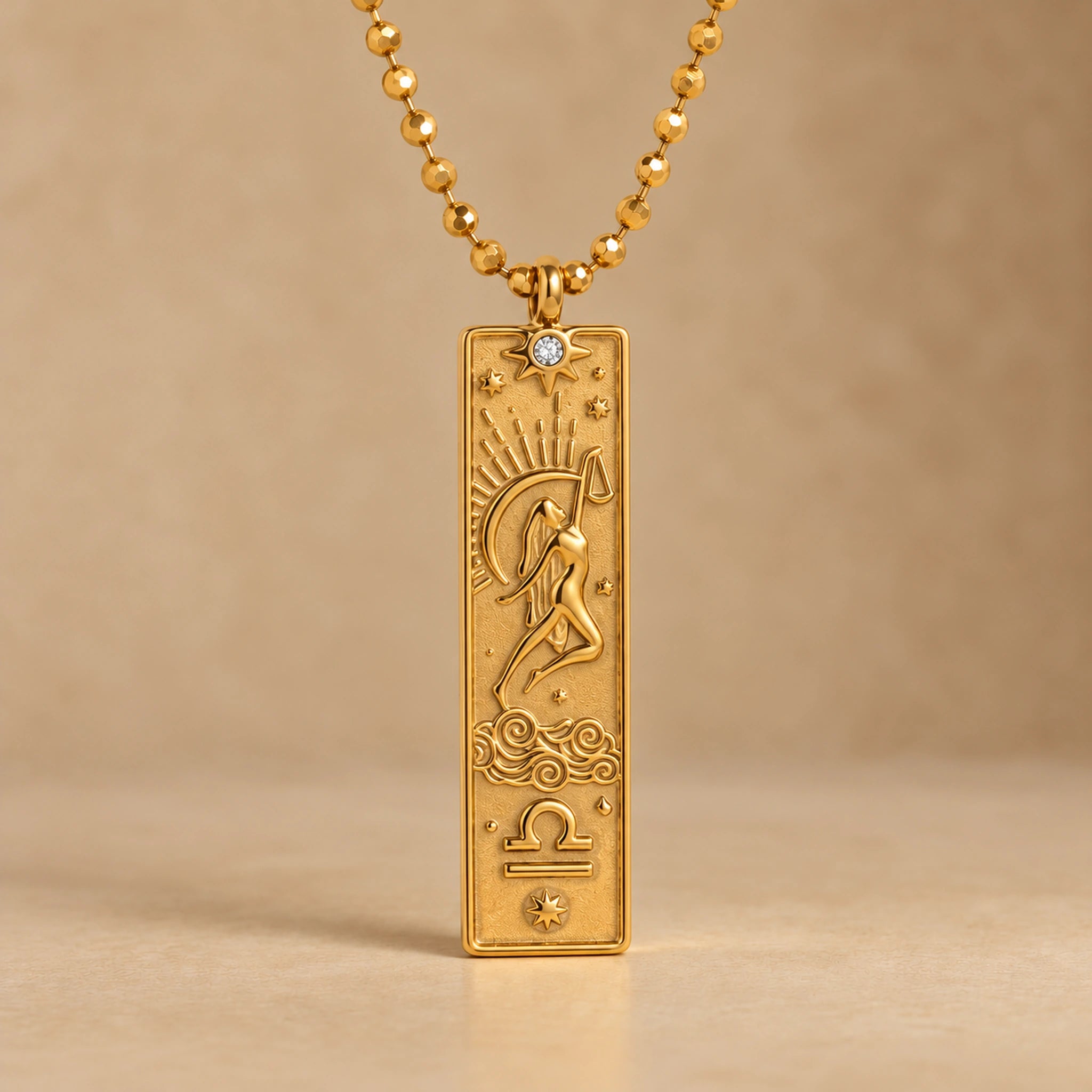 Gold Libra zodiac necklace, embossed bar pendant with sign symbol on a faceted ball chain, warm beige background