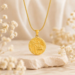 A gold pendant necklace with a circular libra zodiac sign design, resting on a textured stone with soft dried flowers in a warm, neutral setting.