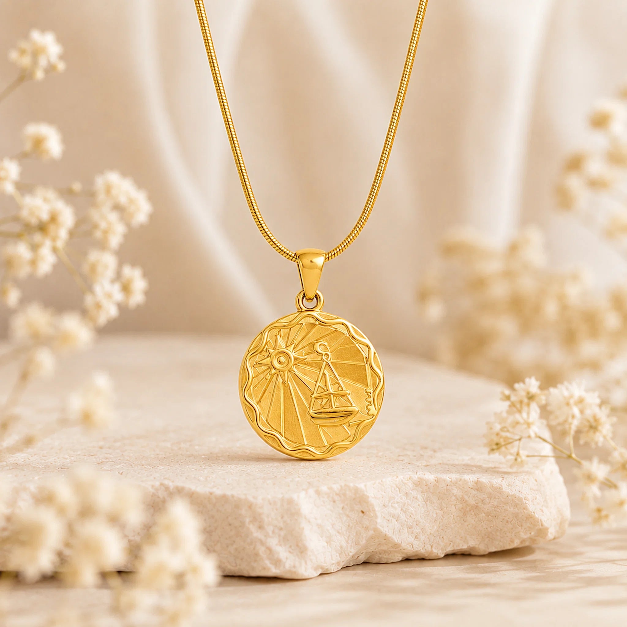A gold pendant necklace with a circular libra zodiac sign design, resting on a textured stone with soft dried flowers in a warm, neutral setting.
