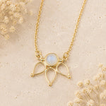 A gold pendant necklace featuring a round iridescent moonstone at the center of an openwork leaf-like design, displayed on a textured beige surface with dried flowers.