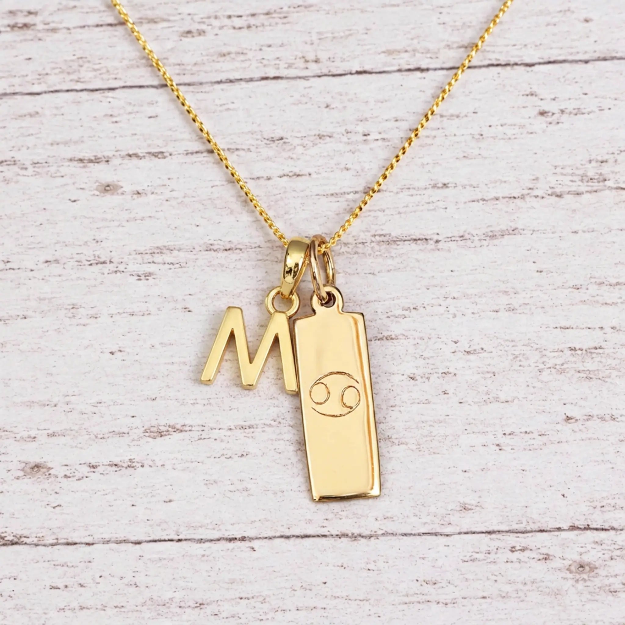 Gold Initial M Pendant and Cancer Zodiac Pendant Necklace on a fine gold chain against a light wooden background