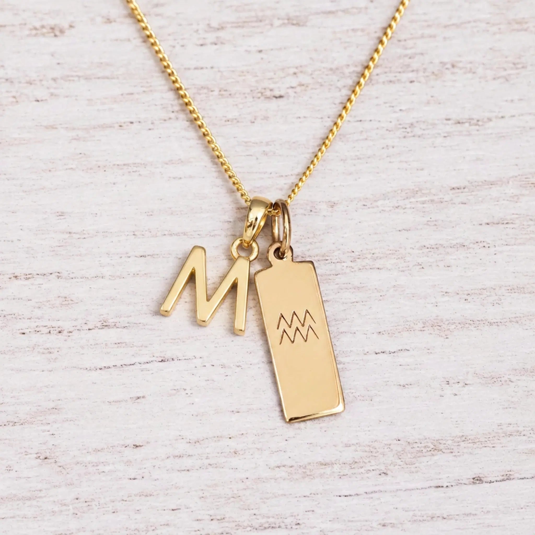 gold M initial and aquarius zodiac necklace on fine gold chain against a light wooden background