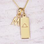 Gold initial and Libra zodiac necklace with a letter M charm and engraved bar pendant on whitewashed wood